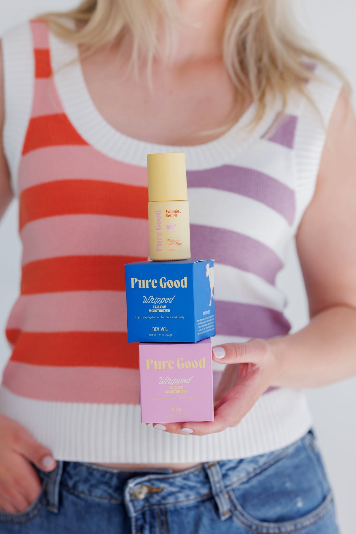 Woman in a striped tank top balancing the Pure Good Vitamin C serum on top of the Pure Good revival tallow blend on top of the Pure tallow blend.
